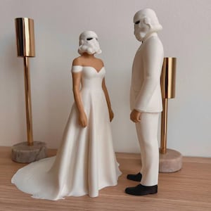 Könnte beinhalten: Hochzeitstortenfiguren mit einer Braut und einem Bräutigam in Weiß, mit Stormtrooper-Helmen. Die Braut trägt ein schulterfreies Kleid. Der Bräutigam trägt einen weißen Anzug. Zwei goldene Kerzenhalter befinden sich auf beiden Seiten.