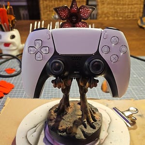 Puede incluir: Un mando de PlayStation 5 blanco y negro pintado a medida, con una base inspirada en el Demogorgon y una cabeza roja en forma de flor. El mando tiene un diseño artístico único, para jugadores y coleccionistas.