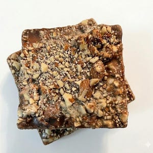 May include: Two square pieces of toffee candy, stacked on top of each other. The toffee is covered in chopped nuts, with a dark brown, glossy base. The candy sits on a white surface.