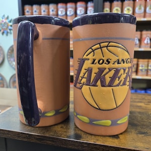 May include: Two ceramic mugs with purple handles and a brown, yellow and purple Los Angeles Lakers basketball design. The mugs are on a dark brown surface.
