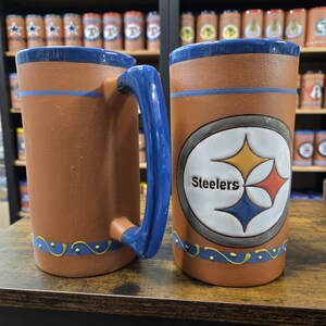 May include: Two brown ceramic mugs with blue handles and blue and yellow trim. One mug features the Pittsburgh Steelers logo with the words "Steelers" in white.
