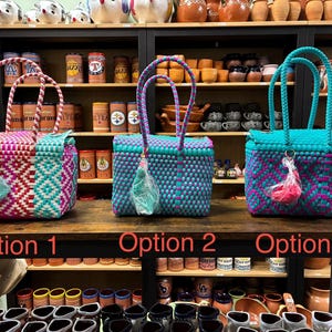 May include: Three rectangular woven plastic baskets with handles, labeled Option 1, Option 2, and Option 3. Each basket features a geometric pattern in vibrant colors including pink, teal, and orange, with a small decorative tassel attached to the front.