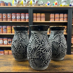May include: Three gray ceramic vases with black floral designs. The vases are round and have a wide mouth. They are sitting on a wooden table.