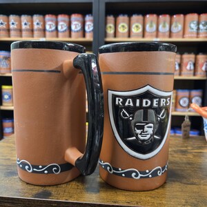 May include: Two brown ceramic mugs with black handles and black trim. One mug features a white Raiders logo with a black helmet and the text "RAIDERS".