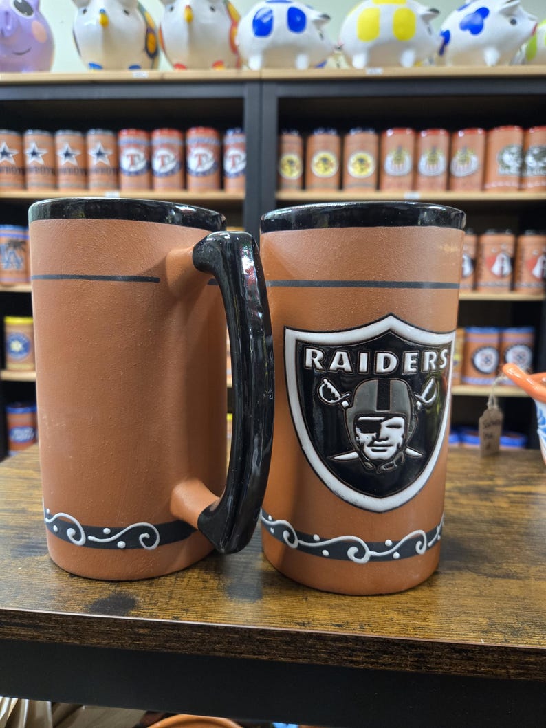 May include: Two brown ceramic mugs with black handles and black trim. One mug features a white Raiders logo with a black helmet and the text "RAIDERS".