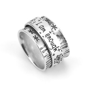 May include: Silver spinning ring with the words "I am enough" engraved on the band. The ring is decorated with small flower designs.