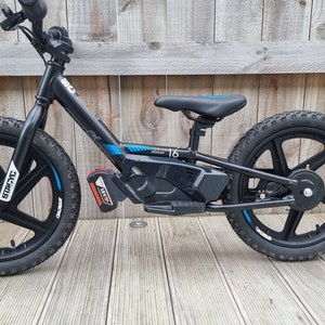 Puede incluir: Bicicleta de equilibrio eléctrica negra con detalles azules y un asiento negro. La bicicleta tiene una batería en el cuadro y ruedas de 40,6 cm.