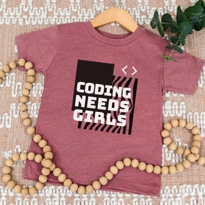 May include: A maroon t-shirt with a black and white graphic that says "Coding Needs Girls" with angle brackets on the right side of the graphic.