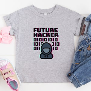 Puede incluir: Camiseta gris con el texto "FUTURE HACKER" en negro y rosa con un patrón de código binario y una imagen de dibujos animados de una persona con una sudadera con capucha.