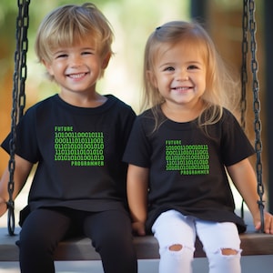 Może przedstawiać: Dwoje dzieci w czarnych koszulkach z napisem "FUTURE 1010011000101111 0001101011001001 0110110100101010 1101010100101010 10101010100101010 PROGRAMMER" w zielonym kolorze.