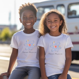 Puede incluir: Dos niños con camisetas blancas con un gráfico de átomo azul y el texto "Future Physicist". Los niños sonríen y miran a la cámara.