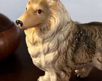 May include: A ceramic figurine of a Collie dog. The dog is standing with its head turned to the side. The figurine is painted in shades of brown, white, and black.