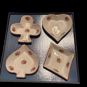 May include: A set of four white porcelain dishes, shaped as a club, heart, spade, and diamond. Each dish features pink rose decorations and gold edging. The dishes are presented in a blue box.