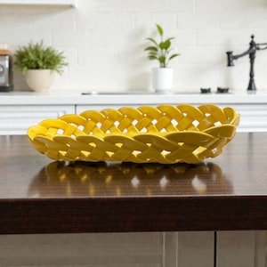 May include: A vibrant yellow ceramic serving basket with a woven design. The oblong dish is placed on a dark brown wooden surface. The kitchen background features a white tiled wall, a plant, and a black tap.