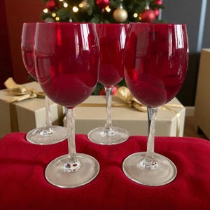 May include: Four ruby red wine glasses with clear stems and bases are displayed on a red cloth. A Christmas tree with gold and red baubles is in the background. Gift boxes with gold ribbon are also visible.