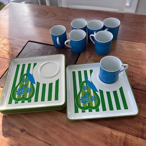 May include: A set of six blue mugs with white interiors and handles, paired with square plates. The plates feature a green and white striped pattern with a pear design in green and blue. The set is arranged on a wooden table.