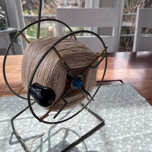 May include: A vintage metal yarn winder featuring a circular frame with a central spool wound with beige thread. The device includes a black handle for rotation, a blue accent on the central axis, and a sturdy rectangular metal base stand.