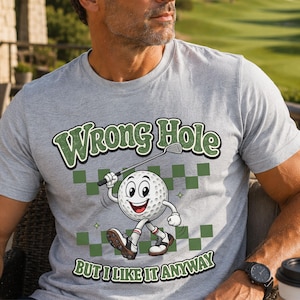 May include: A heather gray t-shirt featuring a cartoon golf ball character holding a golf club. The text reads "WRONG HOLE" at the top and "BUT I LIKE IT ANYWAY" at the bottom, set against a green and gray checkered background pattern.
