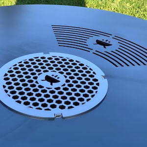 Könnte beinhalten: Nahaufnahme einer runden, silbernen Tischplatte aus Metall mit zwei ausgeschnittenen Designs. Das Design auf der linken Seite ist ein Wabenmuster mit einem Grill-Symbol in der Mitte. Das Design auf der rechten Seite ist eine Reihe von geschwungenen Linien mit einem Grill-Symbol in der Mitte.