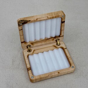 May include: A wooden fly fishing box with a hinged lid and a white foam interior. The box is made of light wood with dark brown streaks.