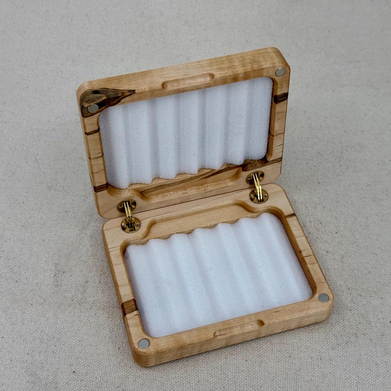 May include: A wooden fly fishing box with a hinged lid and a white foam interior. The box is made of light wood with dark brown streaks.