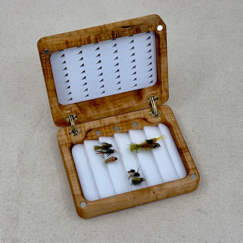 May include: A wooden fly fishing box with a hinged lid and a white foam interior. The box is open and contains several flies.