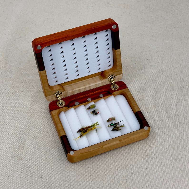 May include: A wooden fly fishing box with a hinged lid. The box is made of maple and walnut wood and has a white foam interior. The box is open and contains several fly fishing lures.