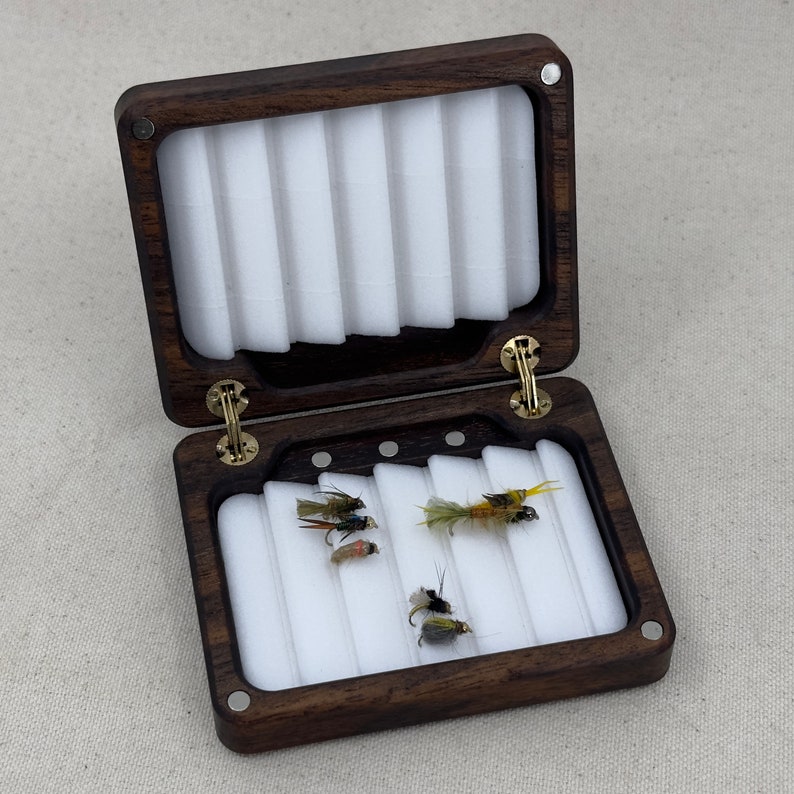 May include: A dark brown wooden fly fishing box with a white foam interior and five compartments. The box is open and contains five flies.