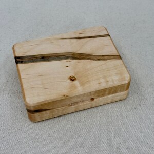 May include: A small wooden box with a natural wood finish and dark brown wood grain accents. The box has a hinged lid and is made of maple wood.