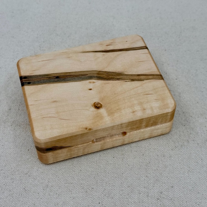 May include: A small wooden box with a natural wood finish and dark brown wood grain accents. The box has a hinged lid and is made of maple wood.