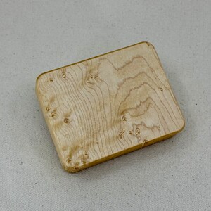 May include: A small, rectangular wooden box with a light brown, natural wood finish. The wood grain is visible and has a unique pattern.