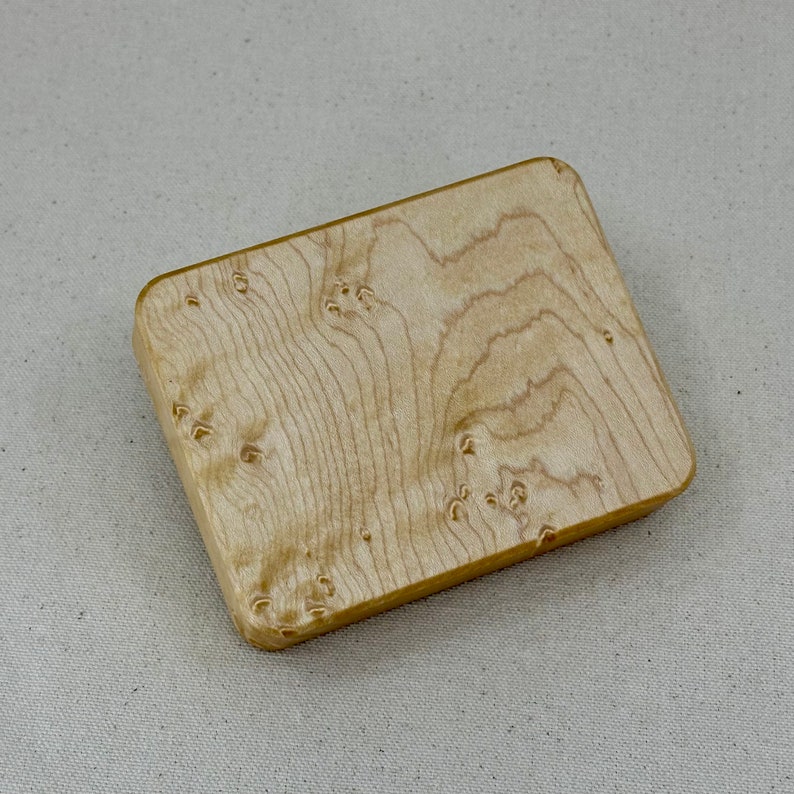 May include: A small, rectangular wooden box with a light brown, natural wood finish. The wood grain is visible and has a unique pattern.
