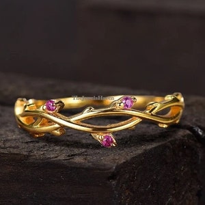 以下が含まれることがあります： 繊細な枝のデザインとピンクの宝石が施された金の指輪。指輪は暗い木製の表面の上に置かれています。
