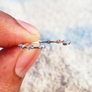Puede incluir: Un anillo fino de metal plateado que presenta una serie de pequeñas piedras redondas y transparentes engastadas a intervalos a lo largo de la banda. El anillo está abierto en la parte superior, mostrando un diseño delicado con piedras brillantes.
