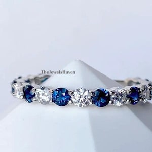 May include: A silver band ring with alternating blue and white round gemstones. The ring is on a white surface. TheJewelsHaven is printed on the surface.