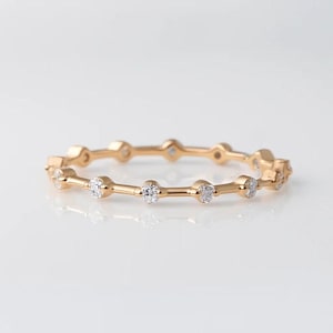 May include: A delicate gold ring with a series of small, round, clear gemstones set along the band. The ring has a minimalist design with a polished finish, reflecting light. The gemstones are evenly spaced, creating a subtle sparkle.