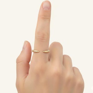 Könnte beinhalten: Ein goldfarbener Ring mit offenem Design. Der Ring hat zwei abgerundete, nicht verbundene Enden, wodurch eine Lücke entsteht. Der Ring wird an einem Finger getragen und unterstreicht seinen minimalistischen und modernen Stil. Der Ring ist wahrscheinlich aus Metall.