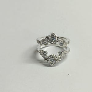 May include: A silver-toned ring guard featuring two symmetrical bands connected at the base. The design includes intricate milgrain detailing and is set with multiple clear, round-cut stones arranged in a pointed, crown-like pattern on each side.