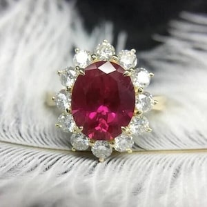 May include: A gold ring featuring a large, oval-cut ruby surrounded by a halo of smaller, round diamonds. The ruby is a deep, vibrant red, and the diamonds sparkle against the gold setting. The ring is set on a white background.