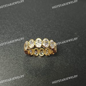 May include: A gold ring with a series of oval-cut, clear gemstones set in a continuous band. The ring is displayed on a dark surface, with the text "HOTSTARJEWELRY" repeated around the edges of the image.