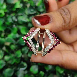 May include: A silver ring featuring a double-layered geometric design. The outer edges are lined with small, round, deep red gemstones, while the inner bands are encrusted with tiny, clear, sparkling white stones, creating a layered look.