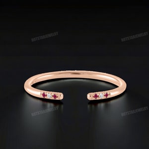May include: A rose gold ring with a split band design. The ring is set with small red and white gemstones.