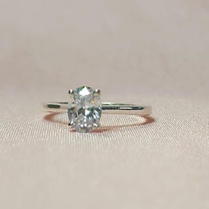May include: A silver engagement ring with a large, oval-cut diamond. The diamond is the focal point, set on a thin, polished band. The ring is displayed against a soft, pink background.