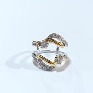 May include: A two-piece bypass ring set crafted from silver-toned metal with gold-toned accents. Each piece features a central round clear stone and a curved band channel-set with small clear stones. The inner band is stamped with 925 hallmark.