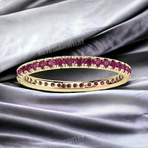 以下が含まれることがあります： 小さな丸い赤い宝石が並んだ金のバンドリング。リングは灰色の布地の背景の上にあります。