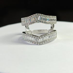 May include: A silver-toned ring guard featuring two chevron-shaped bands connected by vertical bars. Each band is encrusted with a row of baguette-cut stones in the center, bordered by smaller round stones along the top and bottom edges.