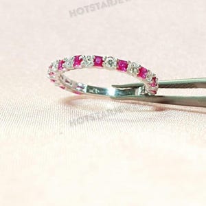 May include: A silver ring with alternating square red gemstones and round clear gemstones. The ring is held by a pair of tweezers against a light pink background. The text "HOTSTARJEWELRY" is visible in the background.