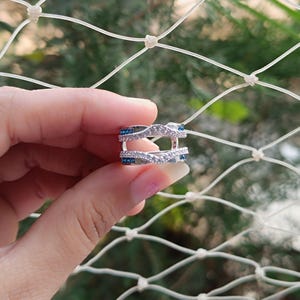 May include: A silver-toned ring guard featuring two chevron-shaped bands connected by a vertical bar. The bands are encrusted with small, clear white stones and accented with small, vibrant blue stones along the sides of the chevron points.
