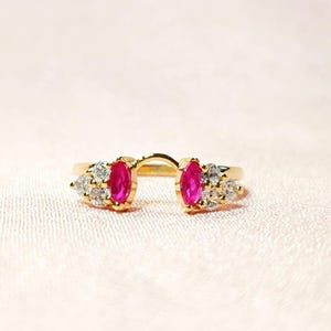 Puede incluir: Un anillo de oro con dos piedras de rubí en forma de marquesa y piedras más pequeñas transparentes a cada lado. El anillo está diseñado para ser usado como un realzador de anillo.