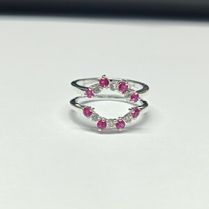 May include: A silver ring guard featuring two curved bands connected at the sides. The bands are set with alternating round-cut vibrant pink stones and clear white stones, creating a symmetrical, open-arched design with a polished metallic finish.